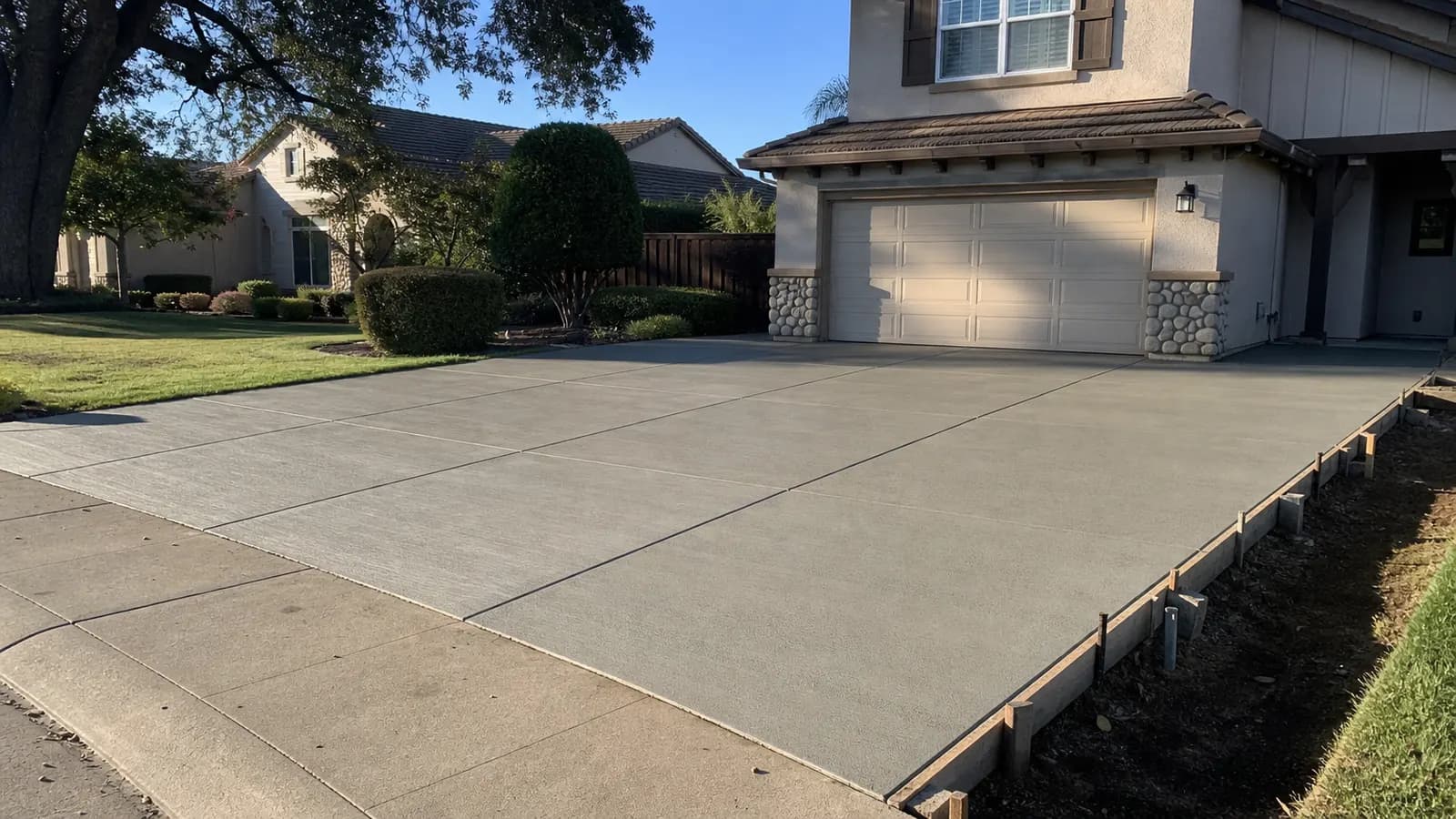 Freshly poured light gray broom-finish concrete driveway in front of a two-story stucco tract home, with saw-cut control joints, form boards along one edge and a valley oak behind